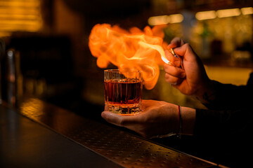 hand of bartender hold old-fashioned glass with alcoholic drink and a piece of ice and make fire near it
