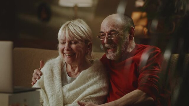 Optimistic elderly couple smiling and waving hands then talking while sitting on couch and making video call to relatives via laptop at home