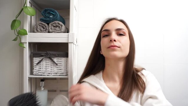 Gorgeous young brunette woman in a bathrobe brushing her hair. Bathroom routine. 