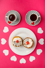 On a pink background, cakes in a plate decorated with paper hearts and two cups of coffee top view.