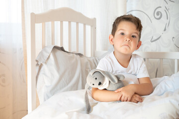 Upset boy in bed at morning. Lonely toddler hugs a toy 