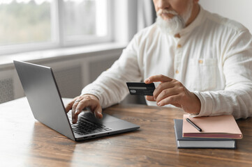 Man pays for an online purchase with a bank card. An old gray man with a beard at the table does online shopping. Close up.