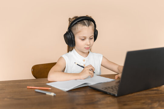 A junior school girl does lessons with a laptop and notebook in large headphones on a beige background.