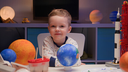 Portrait smiling little blond boy sitting at home at table near planet Earth around many planets of...