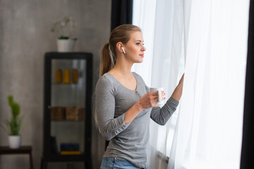 Young teenage girl is standing in front of the window and looking into it. Blond woman at home.