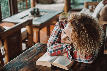 Cute woman asleep on an open book alone at home. Tired female people sleeping on the table. Student concept lifestyle. Cute blonde long curly hair lady resting in living room. Cozy cabin interior