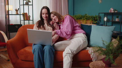 Happy girls friends siblings using laptop computer watching online tv show video sitting on sofa together. Young adult women couple looking at notebook screen making online shopping purchase at home