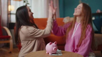 Happy girls friends siblings sitting on floor and take turns dropping dollar banknote into piggy...