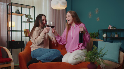 Happy girls friends siblings clinking glasses of red wine cheering and drinking, celebrate birthday party hangout at home in living room. Two female women couple family celebrating new house purchase