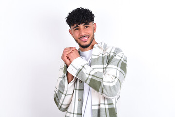 Dreamy young arab man with curly hair wearing overshirt over white background with pleasant expression, closes eyes, keeps hands crossed near face, thinks about something pleasant