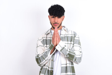 young arab man with curly hair wearing overshirt over white background keeps palms pressed together in front of her having regretful look, asking for forgiveness. Forgive me please.