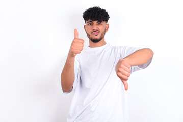 young arab man with curly hair wearing white t-shirt over white background  feeling unsure making good bad sign. Displeased and unimpressed.