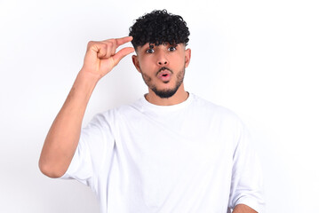 Shocked young arab man with curly hair wearing white t-shirt over white background  shows something little with hands, demonstrates size, opens mouth from surprise. Measurement concept.