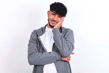 Very bored young arab man with curly hair wearing casual clothes over white background holding hand on cheek while support it with another crossed hand, looking tired and sick.