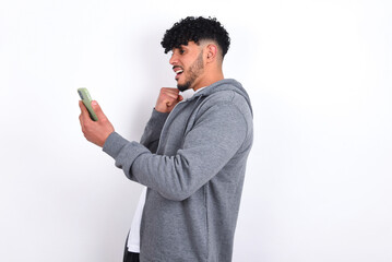 Happy cheerful young arab man with curly hair wearing casual clothes over white background receiving good news via e-mail and celebrating success while standing and looking at mobile phone.