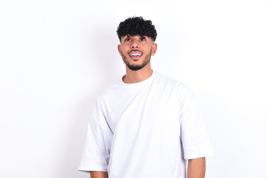 Surprised young arab man with curly hair wearing white t-shirt over white background , shrugs shoulders, looking sideways, being happy and excited. Sudden reactions concept.