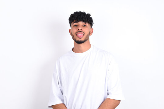 young arab man with curly hair wearing white t-shirt over white background  with happy and funny face smiling and showing tongue.