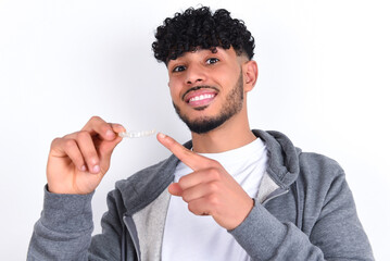 young arab man with curly hair wearing casual clothes over white background holding an invisible aligner and pointing at it. Dental healthcare and confidence concept.