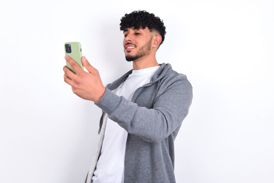 Isolated shot of pleased cheerful young arab man with curly hair wearing casual clothes over white background, makes selfie with mobile phone. People, technology and leisure concept