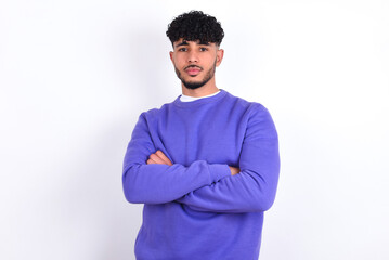 Self confident serious calm young arab man with curly hair wearing purple sweatshirt over white background stands with arms folded. Shows professional vibe stands in assertive pose.