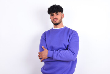 Gloomy dissatisfied young arab man with curly hair wearing purple sweatshirt over white background looks with miserable expression at camera from under forehead, makes unhappy grimace