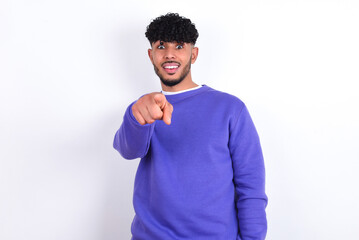 Excited positive young arab man with curly hair wearing purple sweatshirt over white background points index finger directly at you, sees something very funny. Wow, amazing