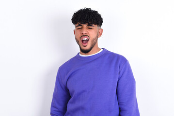 young arab man with curly hair wearing purple sweatshirt over white background yawns with opened...