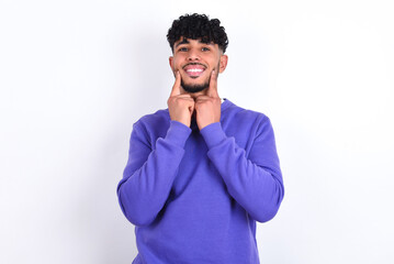 Happy young arab man with curly hair wearing purple sweatshirt over white background with toothy smile, keeps index fingers near mouth, fingers pointing and forcing cheerful smile