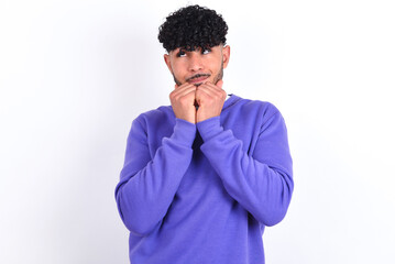 Fototapeta na wymiar Curious young arab man with curly hair wearing purple sweatshirt over white background keeps hands under chin bites lips and looks with interest aside.