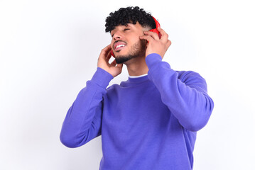 young arab man with curly hair wearing purple sweatshirt over white background with headphones on head, listens to music, enjoying favourite song with closed eyes, holding hands on headset.