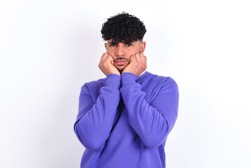 Portrait of sad young arab man with curly hair wearing purple sweatshirt over white background hands face