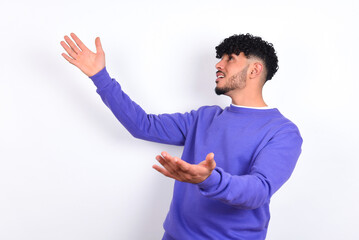 Funny astonished young arab man with curly hair wearing purple sweatshirt over white background look empty space with arms opened ready to catch something.