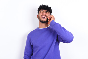 Overemotive happy young arab man with curly hair wearing purple sweatshirt over white background laughs out positively hears funny story from friend during telephone conversation