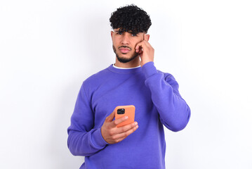 young arab man with curly hair wearing purple sweatshirt over white background holding gadget while sticking out tongue