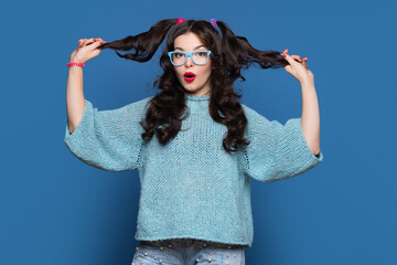 Carefree girl with two ponytails posing on blue background. Young woman wear in casual blue sweater and blue glasses. Make funny faces. Fun and cheerfulness.