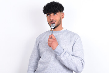 Very hungry young arab man with curly hair wearing sport sweatshirt
over white background holding...
