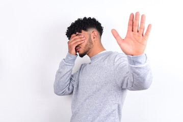 young arab man with curly hair wearing sport sweatshirt
over white background covers eyes with palm and doing stop gesture, tries to hide from everybody.