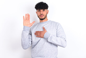 young arab man with curly hair wearing sport sweatshirt
over white background Swearing with hand on...