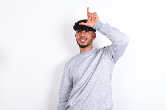 young arab man with curly hair wearing sport sweatshirt
over white background making fun of people with fingers on forehead doing loser gesture mocking and insulting.