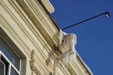 A huge icicle hanging from the roof of an old vintage house next to an open window, a drainpipe and a street led light. Blue sky. - obrazy, fototapety, plakaty