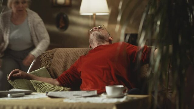 Senior woman hurrying to give tablet to elderly man finishing to read newspaper and falling down on sofa with severe chest pain