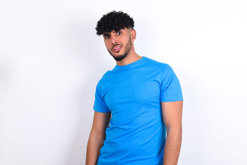 Portrait of dissatisfied young arab man with curly hair wearing blue t-shirt over white background...