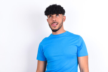 Obraz premium Funny young arab man with curly hair wearing blue t-shirt over white background makes grimace and crosses eyes plays fool has fun alone sticks out tongue.