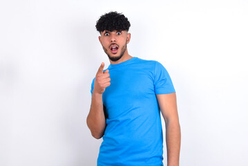 Shocked young arab man with curly hair wearing blue t-shirt over white background points at you with stunned expression