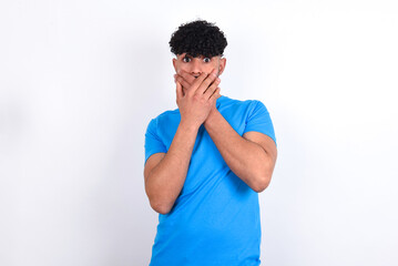 Stunned young arab man with curly hair wearing blue t-shirt over white background covers both hands on mouth, afraids of something astonishing