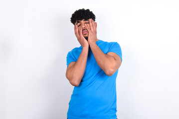 young arab man with curly hair wearing blue t-shirt over white background keeps hands on cheeks has bored displeased expression. Stressed hopeless model