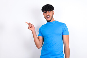 young arab man with curly hair wearing blue t-shirt over white background points aside on copy blank space. People promotion and advertising concept