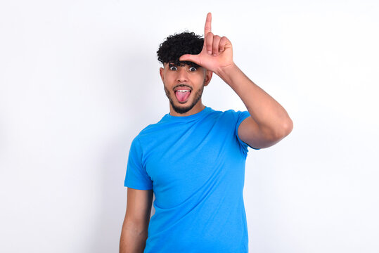 young arab man with curly hair wearing blue t-shirt over white background gestures with finger on forehead makes loser gesture makes fun of people shows tongue
