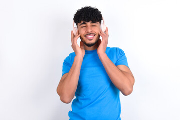 Fototapeta premium Pleased young arab man with curly hair wearing blue t-shirt over white background enjoys listening pleasant melody keeps hands on stereo headphones closes eyes. Spending free time with music
