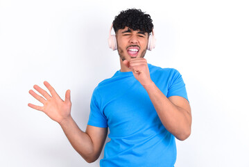 Happy young arab man with curly hair wearing blue t-shirt over white background sings favourite song keeps hand near mouth as if microphone wears wireless headphones, listens music